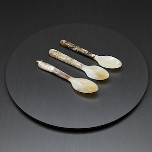 Puede incluir: Tres cucharas decorativas con cuencos de nácar y mangos estampados descansan sobre un plato circular negro. Las cucharas tienen un brillo nacarado, con variaciones de color de crema a marrón.