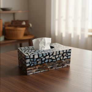 May include: A rectangular tissue box with a mosaic tile design in shades of blue, white, and brown. White tissues are visible extending from the top opening. The box is placed on a wooden surface.