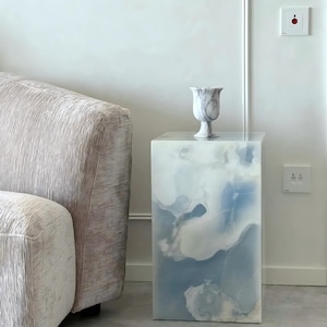Puede incluir: Una mesa pedestal rectangular con un diseño de nubes azules y blancas. Un jarrón de mármol a juego se encuentra encima. La mesa está junto a un sofá texturizado de color claro. La mesa mide unos 50 cm de altura.