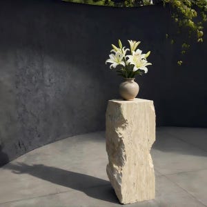 May include: A beige stone plinth supports a small, round, beige vase filled with white lilies. The plinth has a rough, textured surface and is set against a dark grey wall.