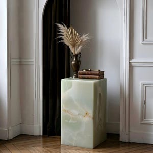 May include: A rectangular, pale green onyx pedestal with a decorative vase and books on top. The vase holds dried flowers and a gold-coloured fan. The pedestal has a marbled appearance and sits on a wooden floor.