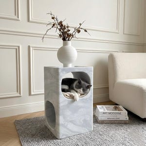 Puede incluir: Una cama para gatos de mármol gris y blanco con un gato durmiendo dentro. La cama tiene una abertura redonda y un jarrón esférico con ramas secas encima. Una pila de libros está al lado de la cama.