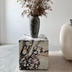 May include: A square marble side table with a gray vase holding dried branches. The table has a white and purple veined pattern. The vase is a dark gray color and has a textured surface. The table is a cube shape.
