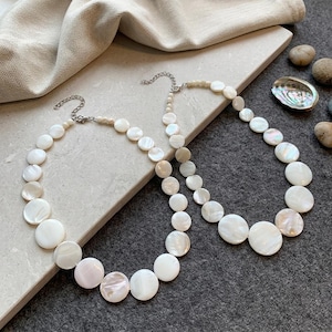 May include: Two necklaces crafted from circular, iridescent white shell beads. Each necklace features a silver-toned metal chain extender with a lobster claw clasp. The beads vary slightly in size and display a natural, pearlescent luster.