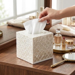 Pode incluir: Uma caixa quadrada para lenços com um mosaico de conchas iridescentes brancas e creme num padrão de escamas. A caixa tem um acabamento liso e refletor com um design de bordos retos, com um lenço branco a sair pela abertura superior.