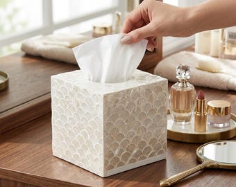 May include: A square tissue box cover featuring a white and cream iridescent shell mosaic in a scalloped pattern. The box has a smooth, reflective finish with a clean, straight-edged design, holding a single white tissue extending from the top opening.