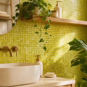 May include: A bathroom vanity area featuring a white oval vessel sink, a brass faucet, and a light wood cabinet. The wall is covered in small, square, lime green glass tiles. A wooden shelf holds a potted plant, a soap dispenser, and a jar.