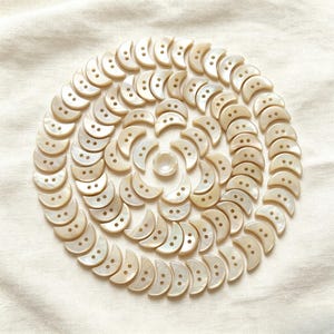 May include: A circular arrangement of off-white, crescent-shaped buttons with two holes each. The buttons are arranged in concentric circles around a single, round button. The background is a cream-colored fabric.