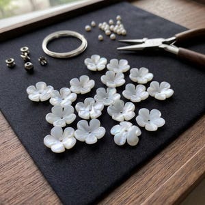 May include: A collection of white mother-of-pearl flower beads, a silver-toned metal ring, small round beads, and metal jewelry pliers. The components are arranged for crafting, featuring iridescent floral shapes with central holes for stringing.