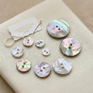 Può includere: Una collezione di otto bottoni rotondi in madreperla iridescente di varie dimensioni, ciascuno con due fori centrali. Sono inclusi una piccola etichetta di carta bianca con il testo Luminous Sea Fasteners Est. 2021 e un sottile ago da cucito.