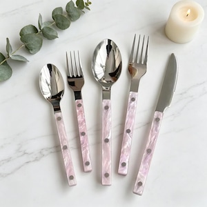 May include: A set of stainless steel cutlery with pink marbled handles. The set includes a spoon, fork, and knife. The handles are accented with silver rivets. The cutlery is arranged on a white marble surface.