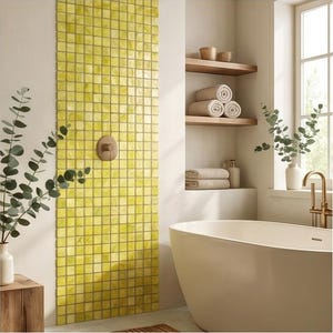 May include: A bathroom interior featuring a vertical accent wall of small, square, yellow mosaic tiles with a circular bronze shower control. A white freestanding bathtub sits nearby, complemented by wooden shelves holding rolled towels and small bowls.