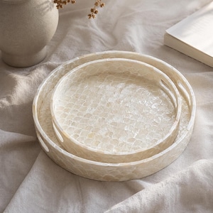 May include: A set of two round nesting trays featuring a shimmering off-white mother-of-pearl inlay arranged in a repeating scalloped pattern. Each tray has a raised edge with integrated cutout handles for easy carrying and a smooth finish.