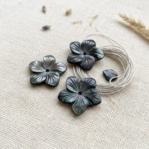 May include: Three dark, iridescent shell buttons carved into a five-petal flower shape with a central hole. The buttons feature a glossy, dark gray finish with subtle green and purple highlights. A coil of thin, light-colored string is included.