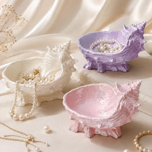 May include: Three seashell-shaped trinket dishes in iridescent white, lavender, and light pink. Each dish features a textured, spiraled shell design with a glossy finish. The white dish holds a pearl necklace, while the lavender dish contains a pearl bracelet.