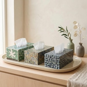 Puede incluir: Tres cajas rectangulares para pañuelos con patrones de mosaico en verde, marrón y gris, cada una con un pañuelo blanco. Las cajas están dispuestas sobre una bandeja larga y ovalada de color crema, junto a dos pequeños jarrones con tallos secos.