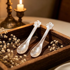 May include: Two white pearlised spoons with flower-shaped handles in a wooden tray. The spoons have a pearlescent sheen and are placed on a dark wooden tray. A lit candle and decorative flowers are in the background.