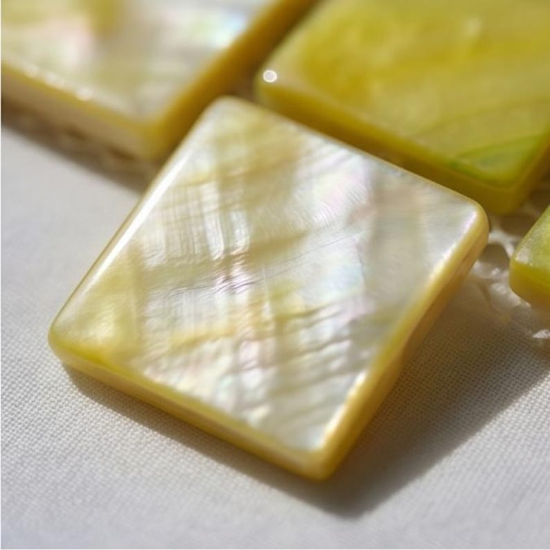 May include: A square button crafted from iridescent mother of pearl with a smooth, polished surface. The material displays a soft yellow hue with natural, shimmering cross-hatched textures and subtle light reflections across its flat face.