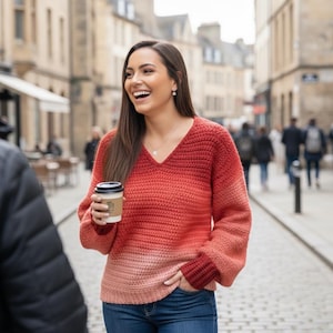 Puede incluir: Una mujer con un suéter de ganchillo ombre rojo y rosa con cuello en V y mangas largas, combinado con jeans azules. Sostiene una taza de café con una funda de papel marrón. El fondo muestra una calle adoquinada y edificios.
