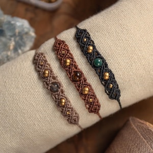 Peut inclure: Trois bracelets en macramé de couleur beige, marron et noir, chacun présentant un motif de cordon tressé avec des perles dorées métalliques intégrées et une perle d'accent plus grande en gris, ambre et vert, présentés sur un rouleau en tissu.