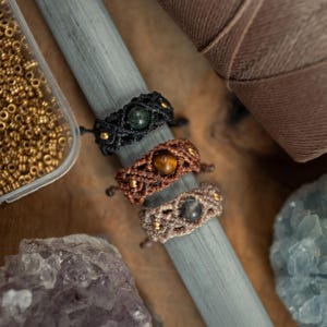 Op de afbeelding: Drie macramé ringen verticaal gerangschikt op een grijze doorn. De bovenste ring is zwart met een donkergroene steen, de middelste is bruin met een tijgeroogsteen en de onderste is beige met een grijze steen. Elke ring heeft kleine gouden kralen.