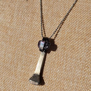 Op de afbeelding: Een zilverkleurige ketting met een spijkerhanger. De spijkerkop heeft een textuur, en de hanger is bevestigd aan een fijne ketting. De spijker is geaccentueerd met een cluster van donkerpaarse kralen. De achtergrond is een beige, getextureerd oppervlak.