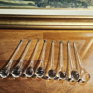 May include: A collection of eight clear glass teardrop ornaments with small metal loops for hanging. The ornaments are displayed on a wooden surface, with a painting in a gold frame in the background. The teardrops are approximately 5 inches long.