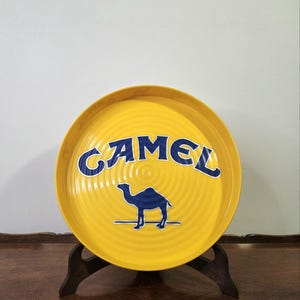 Może przedstawiać: Okrągła żółta taca do serwowania z niebieskim napisem CAMEL oraz grafiką przedstawiającą sylwetkę wielbłąda w kolorze niebieskim na środku. Taca posiada koncentryczną, okrężną fakturę na powierzchni i stoi na ciemnobrązowym drewnianym stojaku.