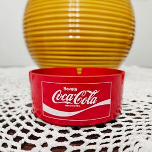 May include: A round red ashtray featuring the white Coca-Cola logo and the text Bevete Coca-Cola MARCHIO REG. The ashtray has two notches on the rim for resting items. The design is centered on the front of the circular red plastic container.
