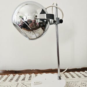 Può includere: Lampada da scrivania vintage cromata con testa sferica e base bianca. La testa della lampada è riflettente. La lampada ha un design minimalista, con stelo cromato e cavo di alimentazione bianco. La base misura circa 15 cm di diametro.
