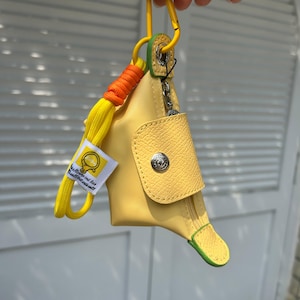 May include: A yellow pouch with a flap closure and a silver button. The pouch has a yellow cord with an orange accent and a yellow tag with text. A yellow carabiner clip is attached to the top.