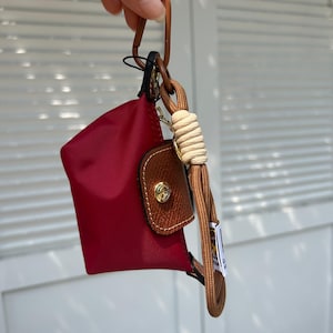 May include: A small, red pouch with a brown leather flap and gold-tone hardware. It hangs from a brown cord and a tan rope loop. The pouch is attached to a brown metal clasp.