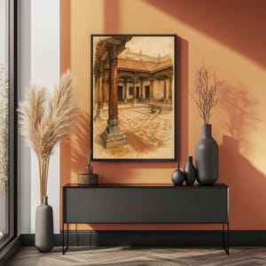 May include: A framed wall art print depicting a watercolor painting of an open-air courtyard with stone flooring, wooden pillars, and a tiled roof. The artwork is displayed in a thin black frame, centered above a long, dark gray console table.
