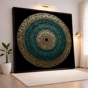 May include: Large framed artwork featuring a circular design of Arabic calligraphy in teal and gold against a black background. The artwork is displayed on a white wall, with a small plant and a rug in the foreground.