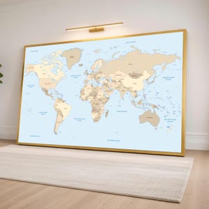 Può includere: Una grande mappa del mondo con cornice dorata, raffigurante continenti e oceani in tonalità beige e blu. La mappa è montata su una parete sopra un tappeto neutro, con un'apparecchio di illuminazione dorato sopra. Un elemento decorativo.