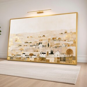 May include: A large rectangular landscape painting in a gold frame featuring a hillside village with white houses, dark trees, and textured gold leaf accents against a neutral beige background, illuminated by a wall-mounted light fixture above.