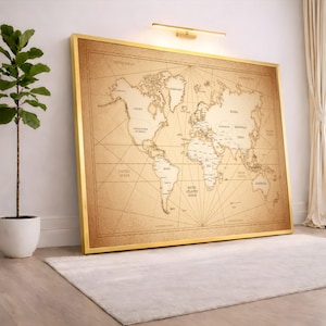 May include: A large world map print with a gold frame, leaning against a white wall. The map is in a vintage style with beige and brown tones, and features country names and ocean labels. A plant in a white pot is to the left.