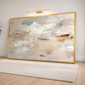 May include: A large rectangular abstract painting featuring thick, textured brushstrokes in shades of beige, cream, white, soft grey, and muted brown. The artwork is enclosed in a thin, metallic gold frame and is mounted on a plain wall.