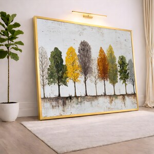 Peut inclure: Peinture de paysage encadrée représentant une rangée d'arbres aux couleurs variées, dont le vert, le jaune et l'orange, sur fond blanc. L'œuvre est dans un cadre doré et est posée contre un mur blanc. Une plante en pot blanc est à gauche.