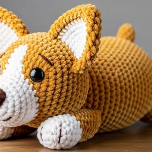 Puede incluir: Un peluche de perro corgi tejido a ganchillo. El perro tiene un cuerpo marrón, detalles blancos en las orejas, el hocico y las patas, y una nariz marrón. Los ojos son negros. El peluche está sobre una superficie de madera.