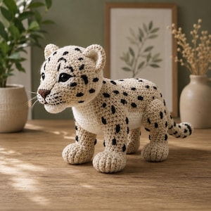 Könnte beinhalten: Ein gehäkeltes Stofftier in Form eines Leopardenbabys aus beigem Garn mit schwarzen Flecken, einem weißen Bauch, einer braunen Nase und schwarzen Kunststoffaugen. Das Spielzeug steht auf vier Beinen mit einem langen Schwanz und zeigt detaillierte Maschen.