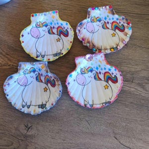 May include: Four decorated seashells featuring a cartoon unicorn design. Each shell has a white base with a unicorn illustration, a rainbow mane and tail, and a star. The edges are adorned with colorful gems.