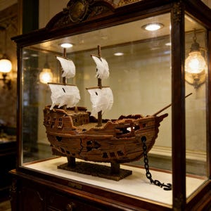 Puede incluir: Maqueta detallada de un barco de madera con velas blancas, expuesta dentro de una vitrina. El barco tiene un aspecto desgastado con daños visibles y está encadenado a la base. La vitrina está colocada sobre un mueble de madera oscura.