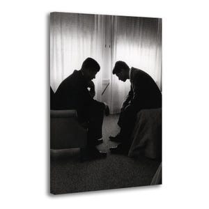May include: Black and white photograph of two men in suits, seated and deep in thought. The men are positioned in front of a window with sheer curtains, creating a sense of privacy and introspection. The image is a study in contrasts.