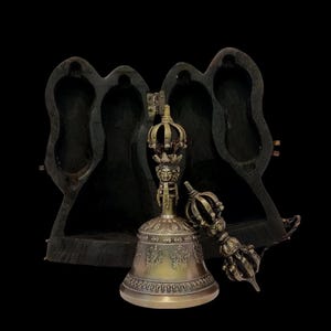 Op de afbeelding: Een koperen rituele bel met een versierd handvat met een gezicht en een kroonvormige top, vergezeld van een bijpassende koperen vajra. Beide items staan voor een donkere, uitgesneden houten kist met twee open, gevormde compartimenten.