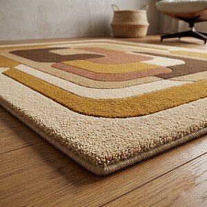 May include: A rectangular area rug featuring a geometric pattern of concentric rounded shapes in shades of cream, mustard yellow, tan, and dark brown. The rug has a thick, textured pile and is finished with a matching bound edge on all sides.