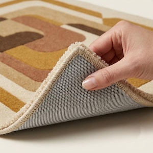 May include: A rectangular area rug featuring a geometric pattern of curved lines in shades of mustard yellow, terracotta, and dark brown on a cream background. The rug has a thick, textured pile and a gray, non-slip, grid-patterned backing.