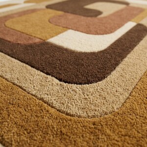May include: A rectangular doormat featuring a textured, tufted surface with a geometric pattern of concentric rounded shapes in shades of dark brown, medium brown, tan, and cream, creating a layered, multi-colored visual effect.