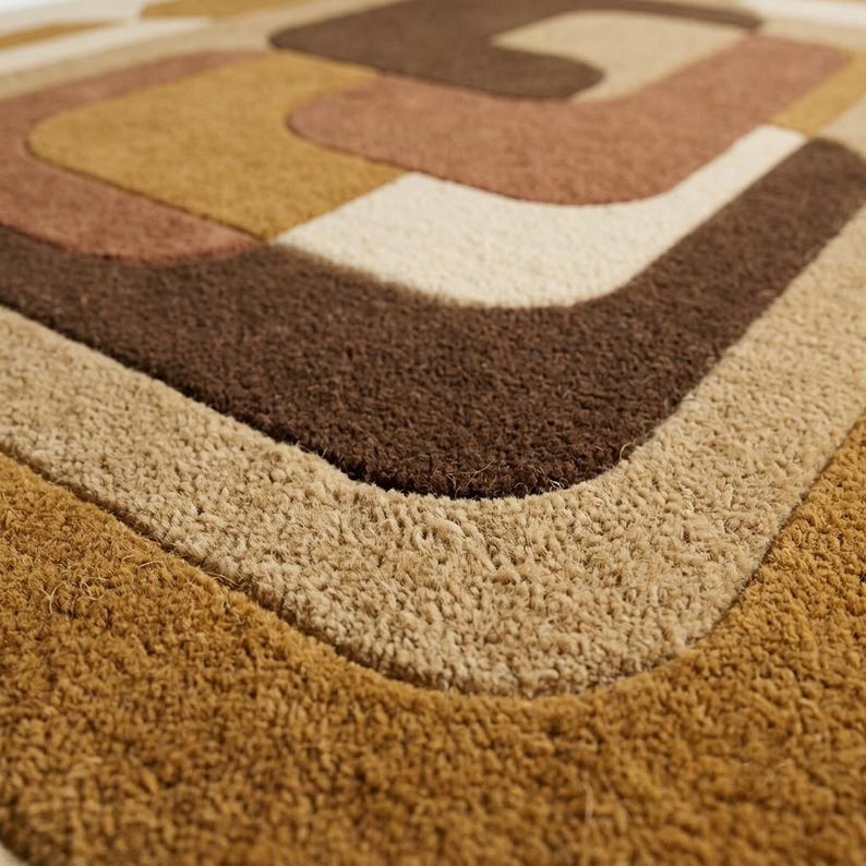 May include: A rectangular doormat featuring a textured, tufted surface with a geometric pattern of concentric rounded shapes in shades of dark brown, medium brown, tan, and cream, creating a layered, multi-colored visual effect.