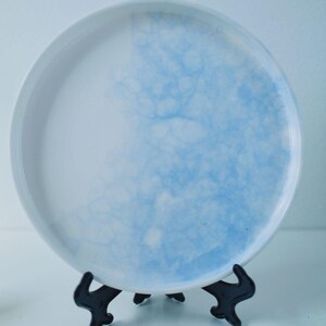 May include: A white ceramic plate with a light blue watercolor design. The plate is displayed on a black stand, showcasing the artistic blue pattern that covers half of the plate's surface.